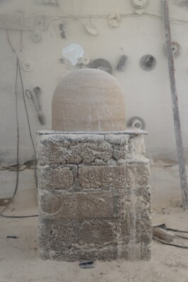 Stone object shaped like a dome sits on a rectangular stone base in a dusty workshop with various tools hanging on the wall in the background. Anomie Publishing