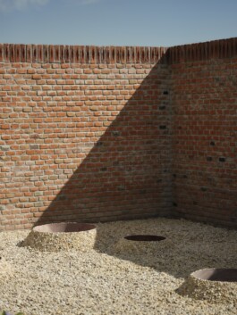 An installation view of the work 'Uketamo' (2024) by Adeline de Monseignat, which is an exterior installation of a brick wall surrounding an area of gravel with three circular holes. Anomie Publishing